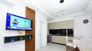 A modern living room with a flat-screen TV, a wooden accent wall, a kitchen with a black marble backsplash, and a dining area with a white table and chairs. - Vila 42 (Vlore)