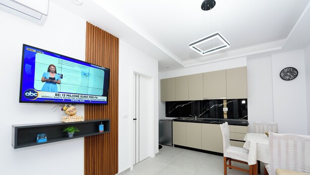A modern living room with a flat-screen TV, a wooden accent wall, a kitchen with a black marble backsplash, and a dining area with a white table and chairs. - Vila 42 (Vlore)