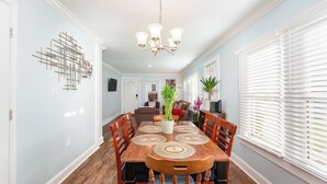 A dining area with a wooden table, chairs, and a chandelier. There are potted plants and a view of a living room in the background. - Modern And Cozy House In The Heart Of Hillsborough (Hillsborough)