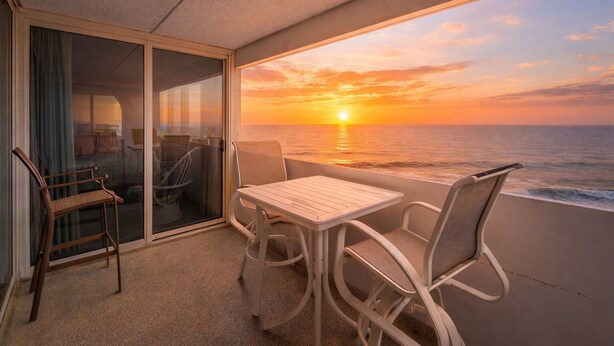 A balcony with a table and chairs, a view of the sunset over the ocean, and a glimpse of the interior through a glass door. - 2BR Atlantis, Family Friendly, 16th fl Oceanfront Views, Outdoor Oceanfront Pool (Ocean City)