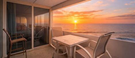 Ein Balkon mit Tisch und Stühlen, Blick auf den Sonnenuntergang über dem Meer und ein Einblick in den Innenraum durch eine Glastür.