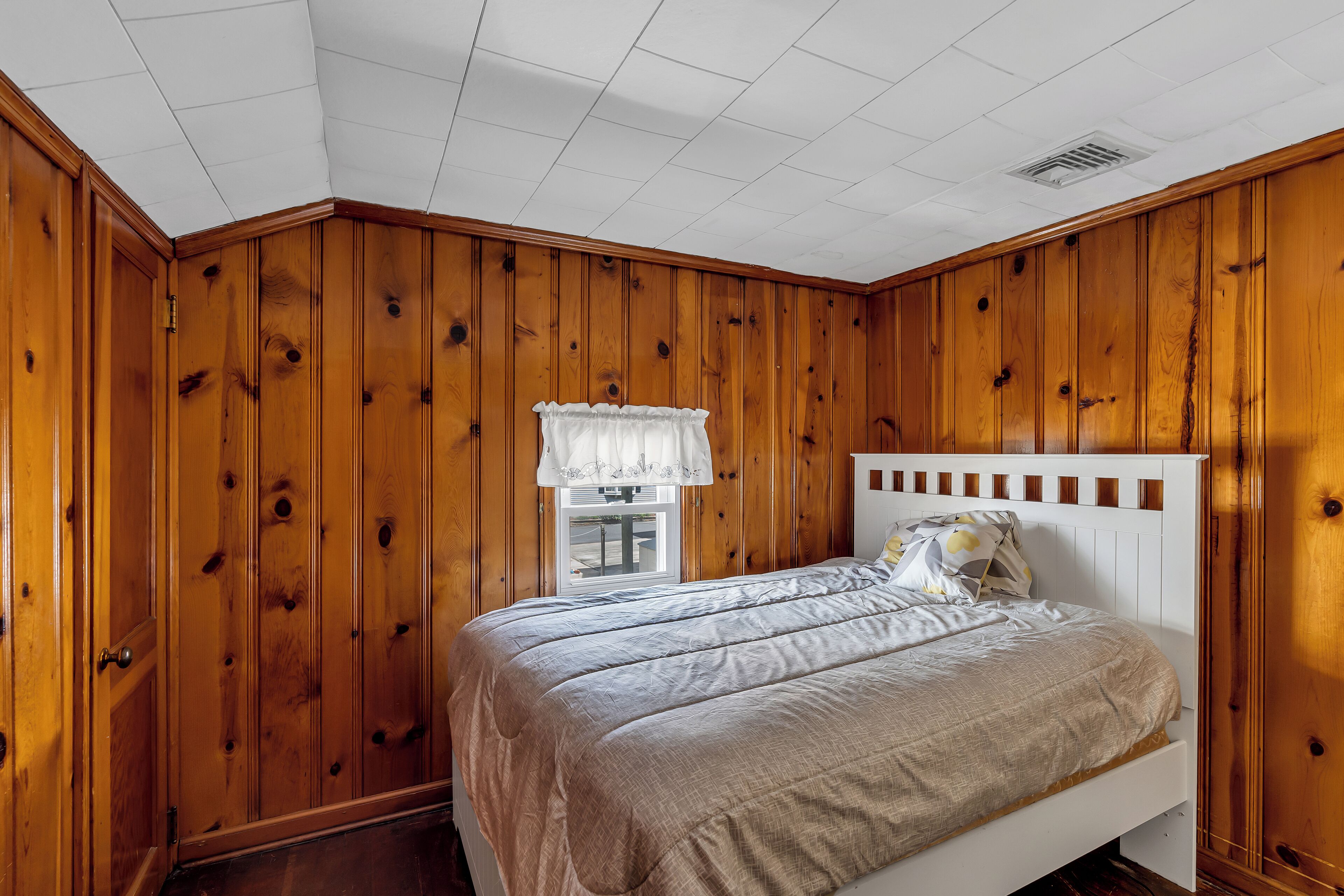 Een slaapkamer met houten lambrisering, een wit bed met hoofdeinde, en een raam met een witte gordijn.