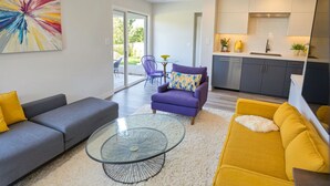 Un salon moderne avec un canapé gris, un canapé jaune et une table basse en verre. On y trouve un fauteuil violet agrémenté d’un coussin coloré. Un grand tableau abstrait orne le mur.