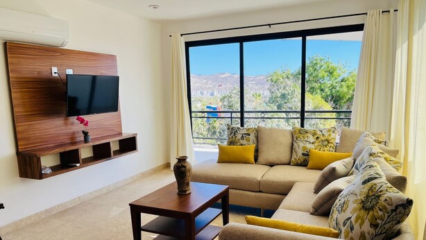 Uma sala de estar moderna com uma televisão grande fixada em uma parede de madeira, um sofá com almofadas florais, uma mesinha de centro e vista para um horizonte urbano através de janelas amplas.