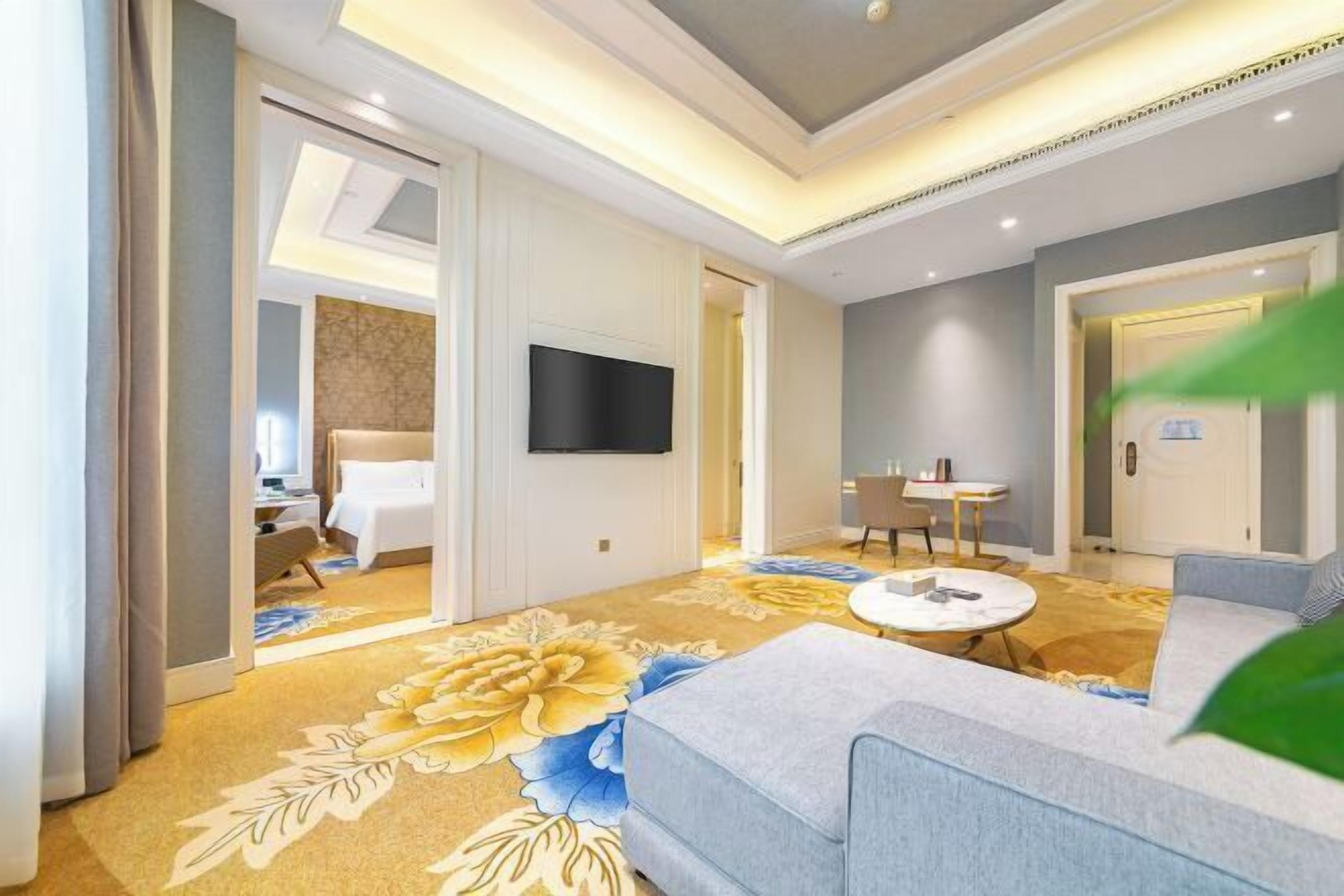 A hotel room with a sofa, a TV mounted on the wall, a dining area with a table and chairs, and a carpet with a floral pattern.