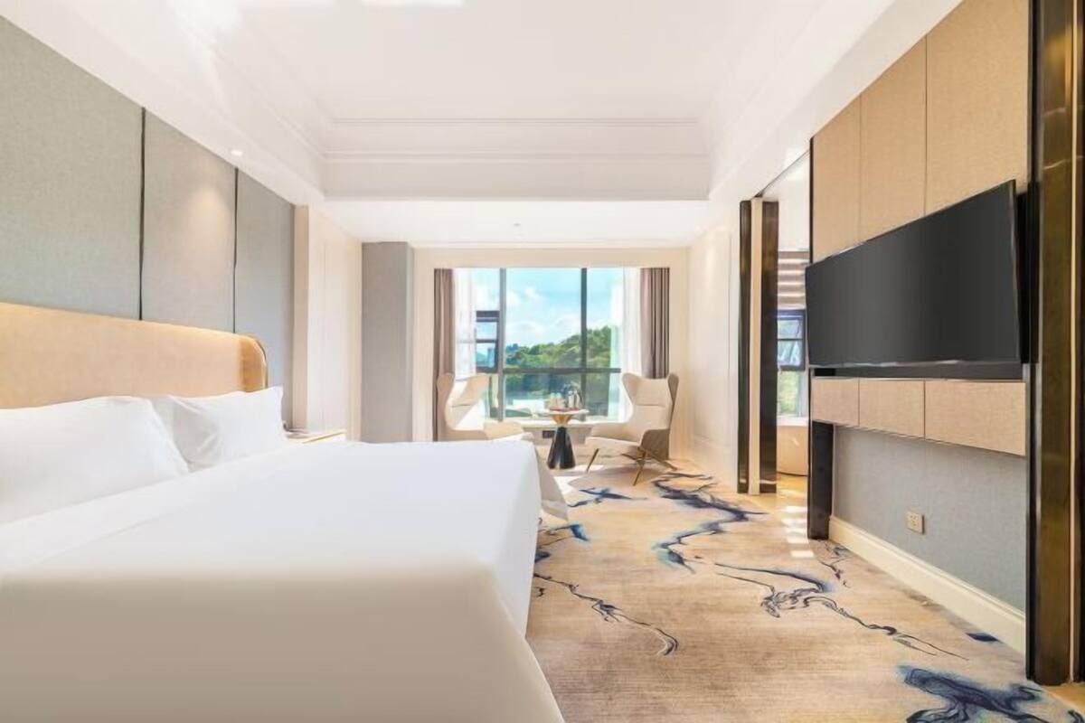 A hotel room with a large bed, a flat-screen TV, a view of the outdoors, and a dining area with chairs.