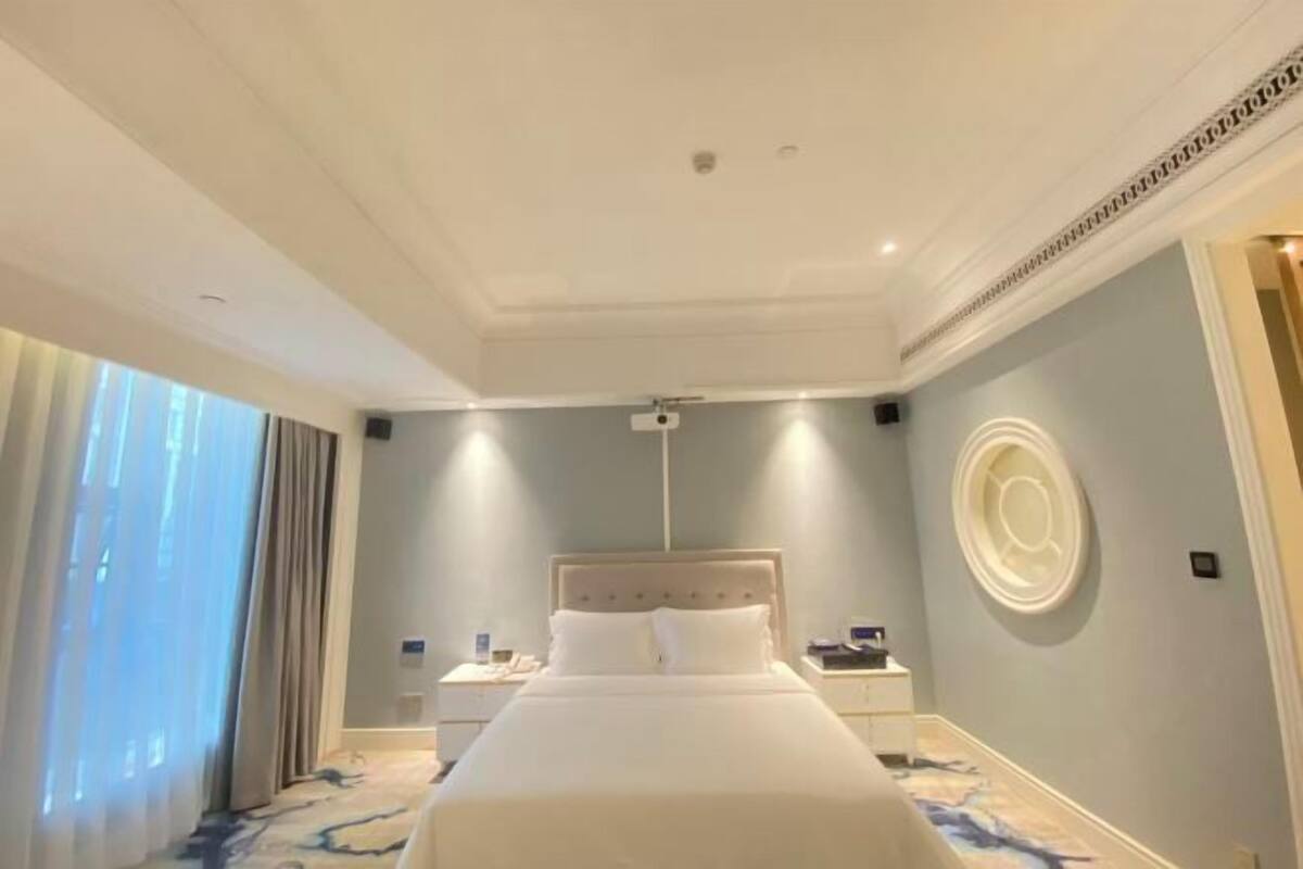 A hotel room with a large bed, a nightstand, a wall clock, and a window with curtains.