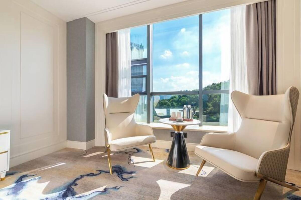 A modern hotel room with two white armchairs, a round black coffee table, and a large window offering a city view.