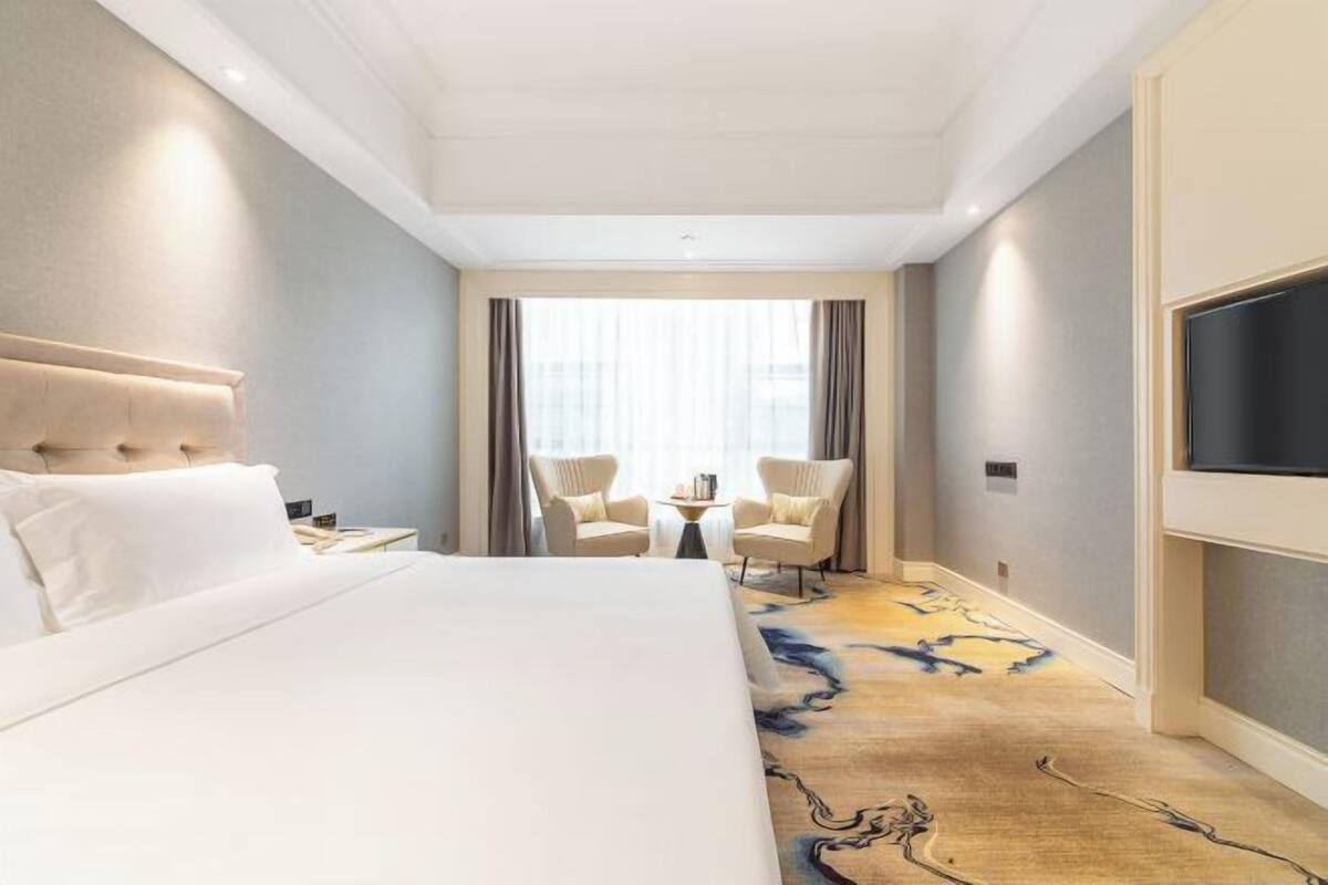 A hotel room with a bed, two armchairs, a small table, a TV, and a large window with curtains.
