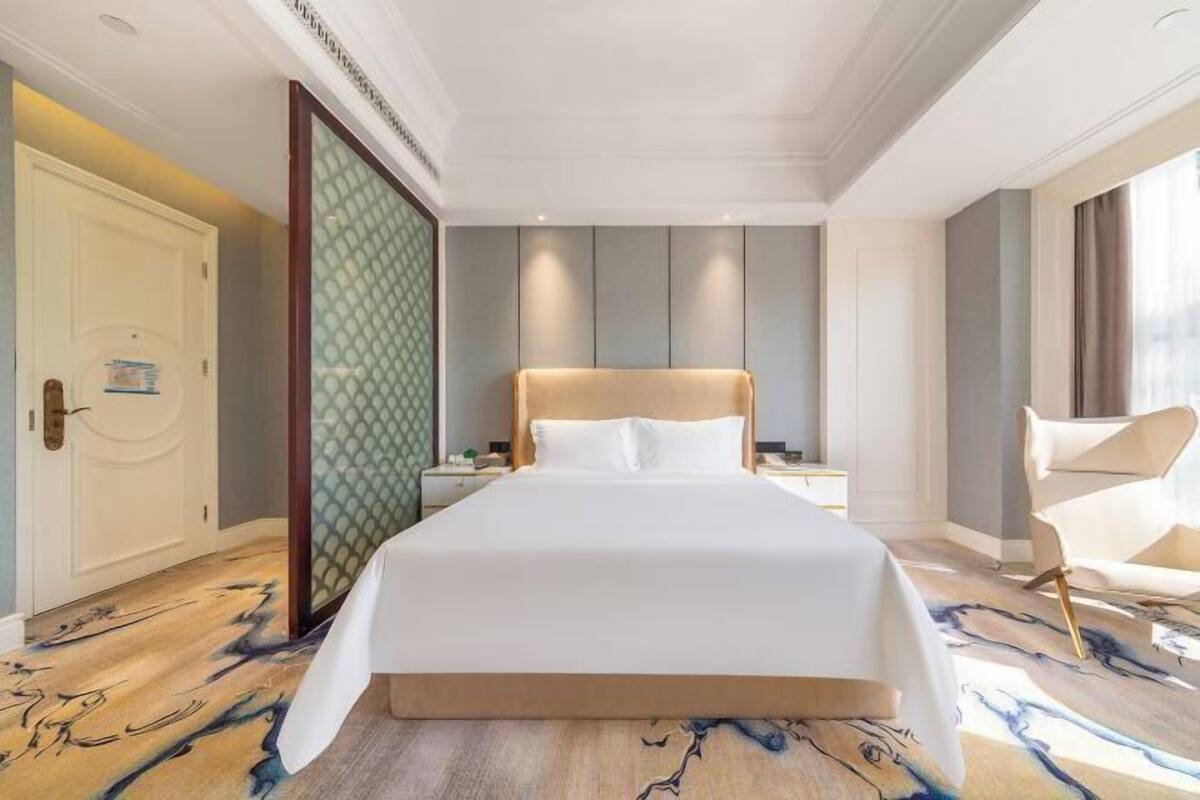 A hotel room with a large bed, a nightstand, a chair, and a view of the outside.