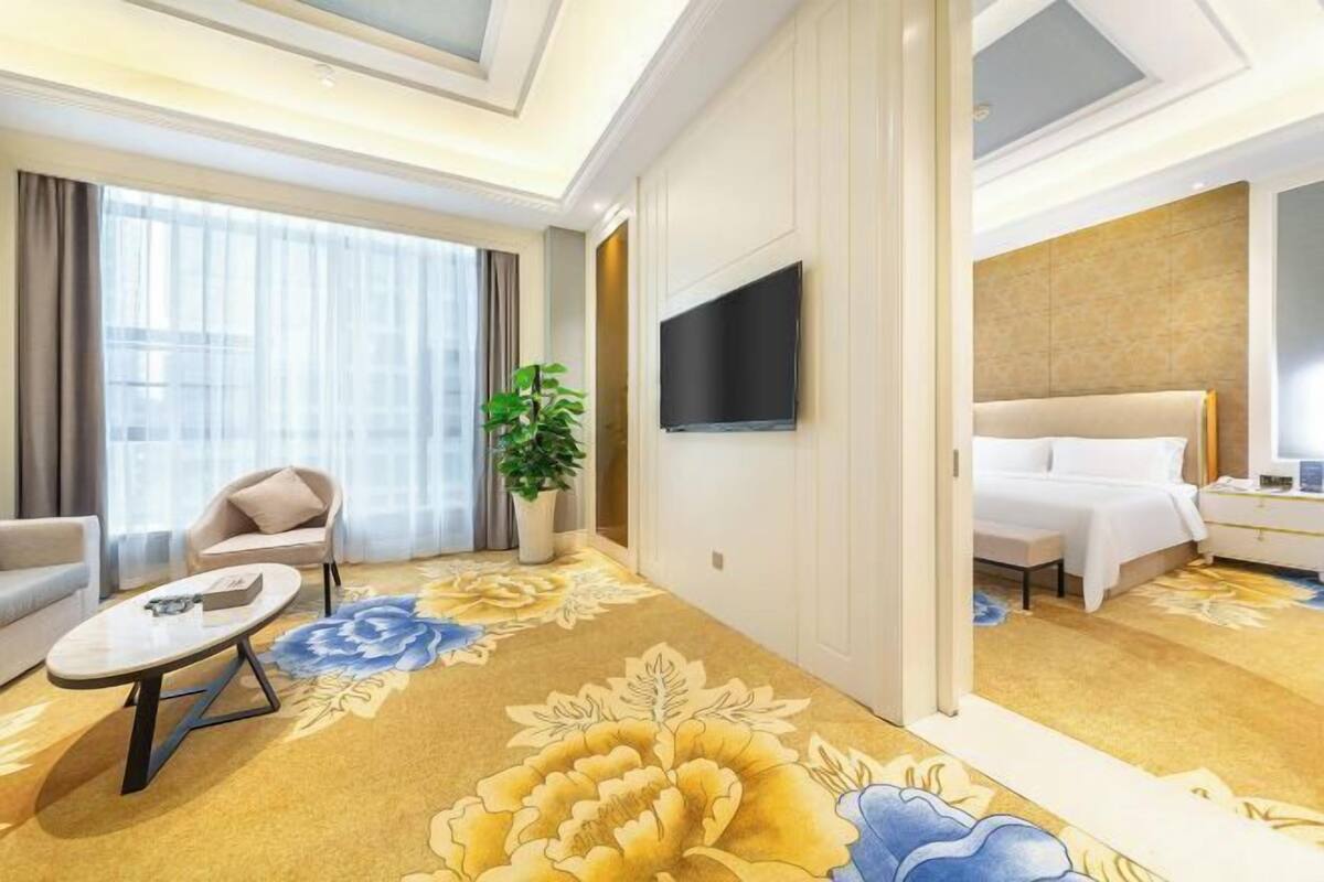 A hotel room with a bed, a TV, a sofa, a coffee table, and a large floral carpet.