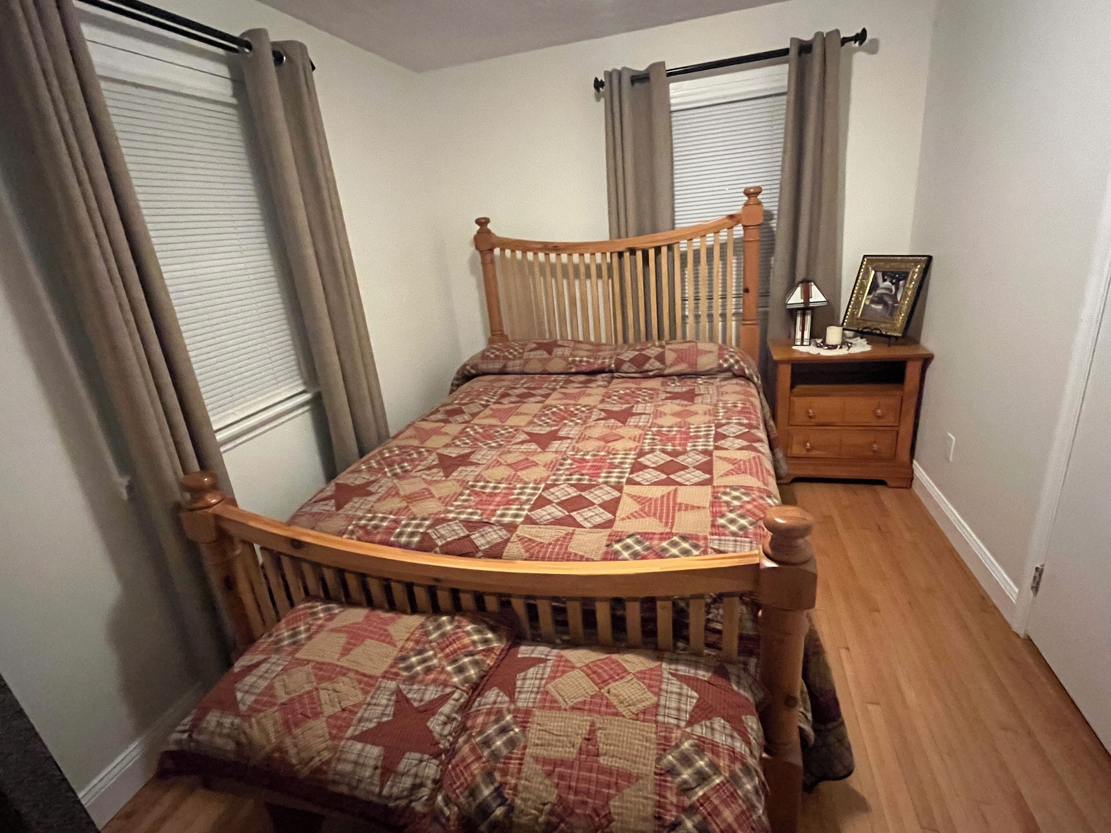 A single bed with a wooden frame and a quilted bedspread, a wooden nightstand with a lamp, and a framed picture.