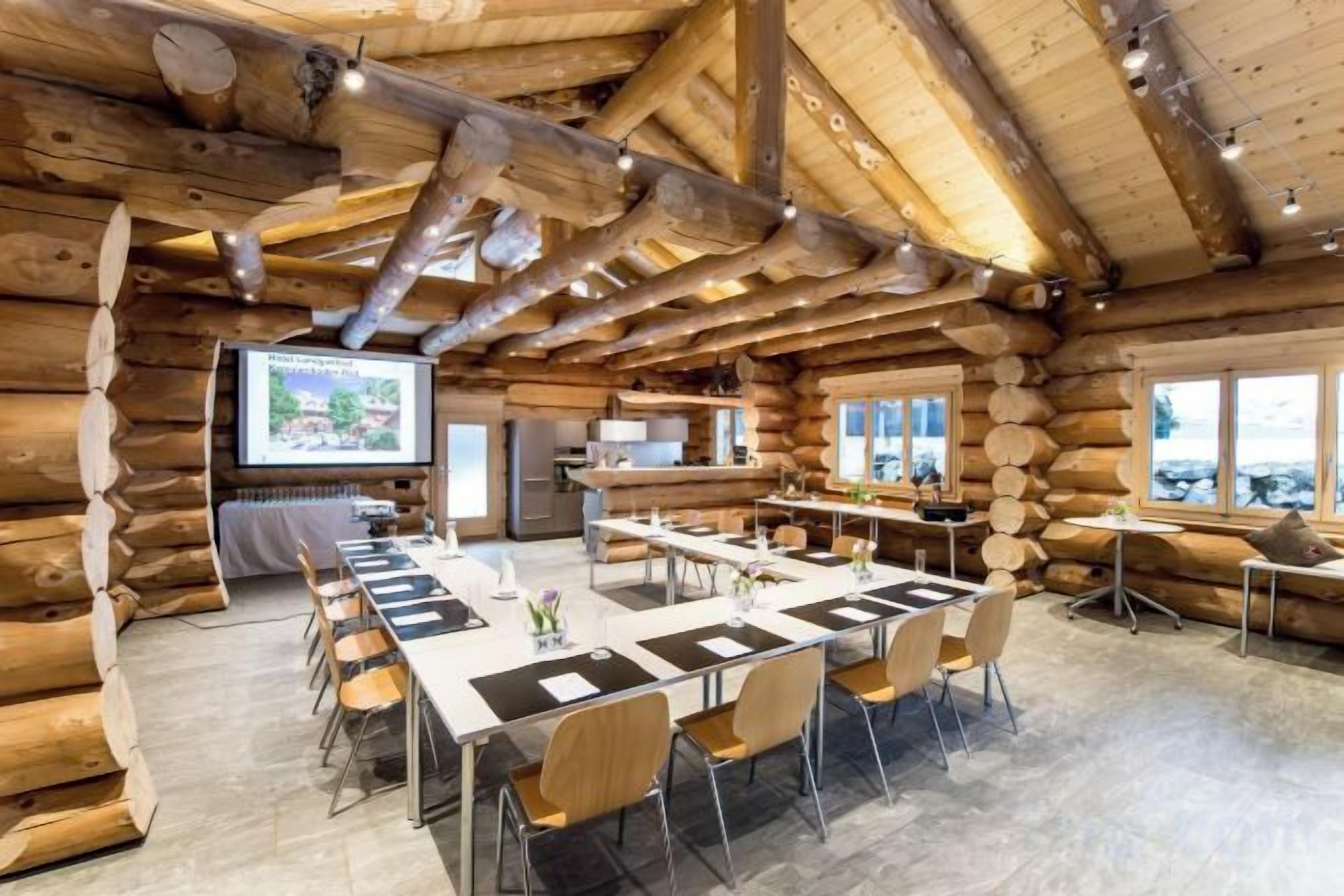 A log cabin-style room with a long table and chairs, a kitchen area, and a large window.