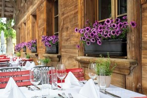 Un restaurante con terraza, mesas dispuestas con manteles blancos y flores moradas en macetas colgantes.