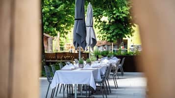 Een buitenterras met tafels, stoelen, parasols en een gebouw op de achtergrond.