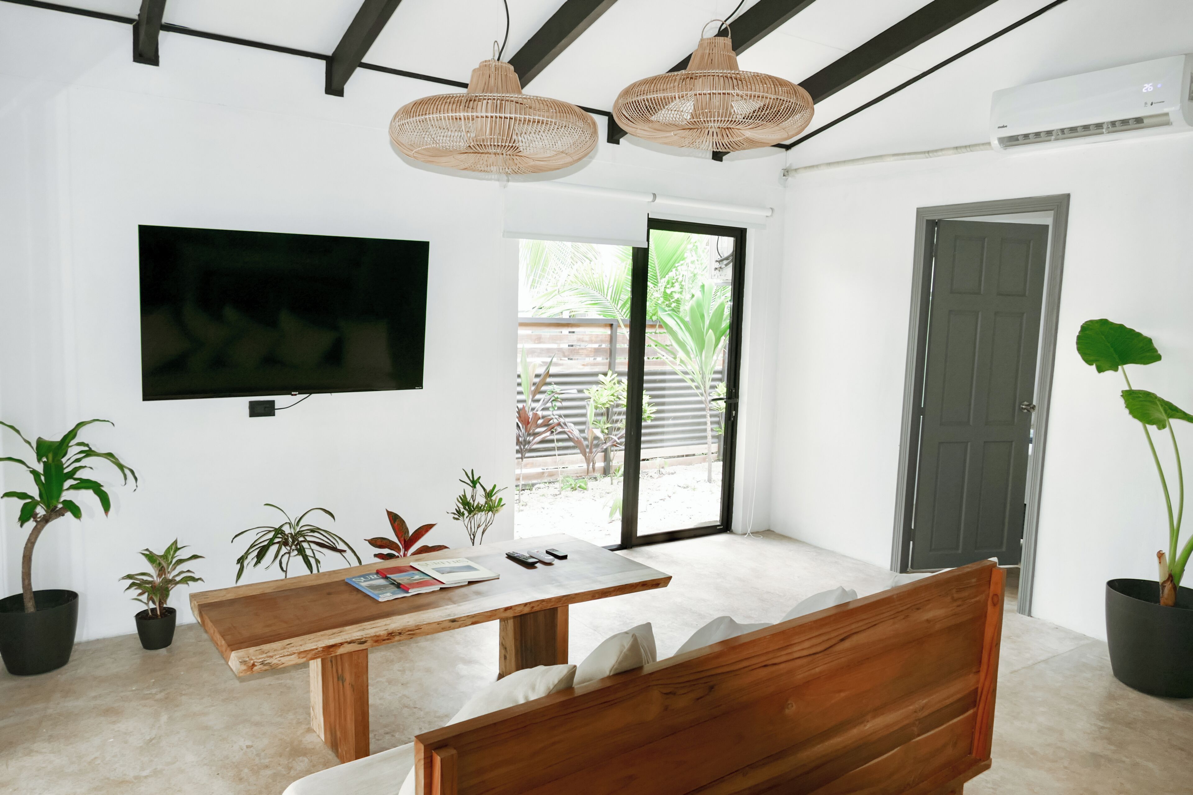 Un salon moderne avec un banc en bois, une table, un téléviseur à écran plat, des luminaires suspendus en osier et des plantes en pot.