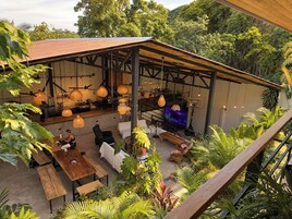 Un área de estar al aire libre con mesas y sillas, rodeada de abundante vegetación y una estructura de pérgola de madera.