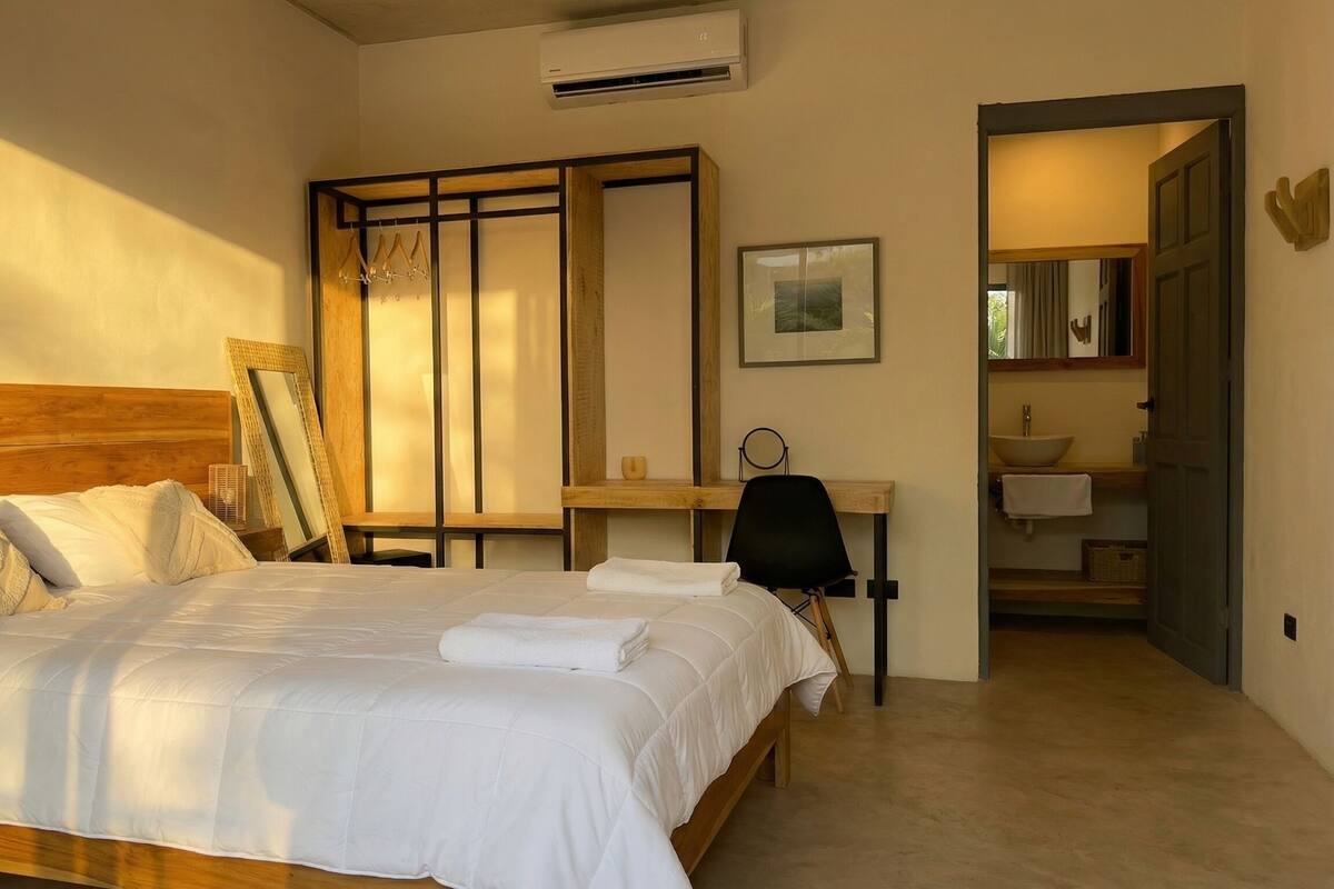 A bedroom with a wooden bed, a desk, a chair, a mirror, and a bathroom visible through an open door.