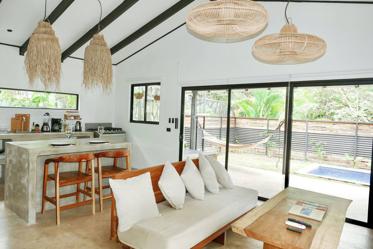 A modern kitchen with a bar area, a sofa, and a dining table with remote controls.