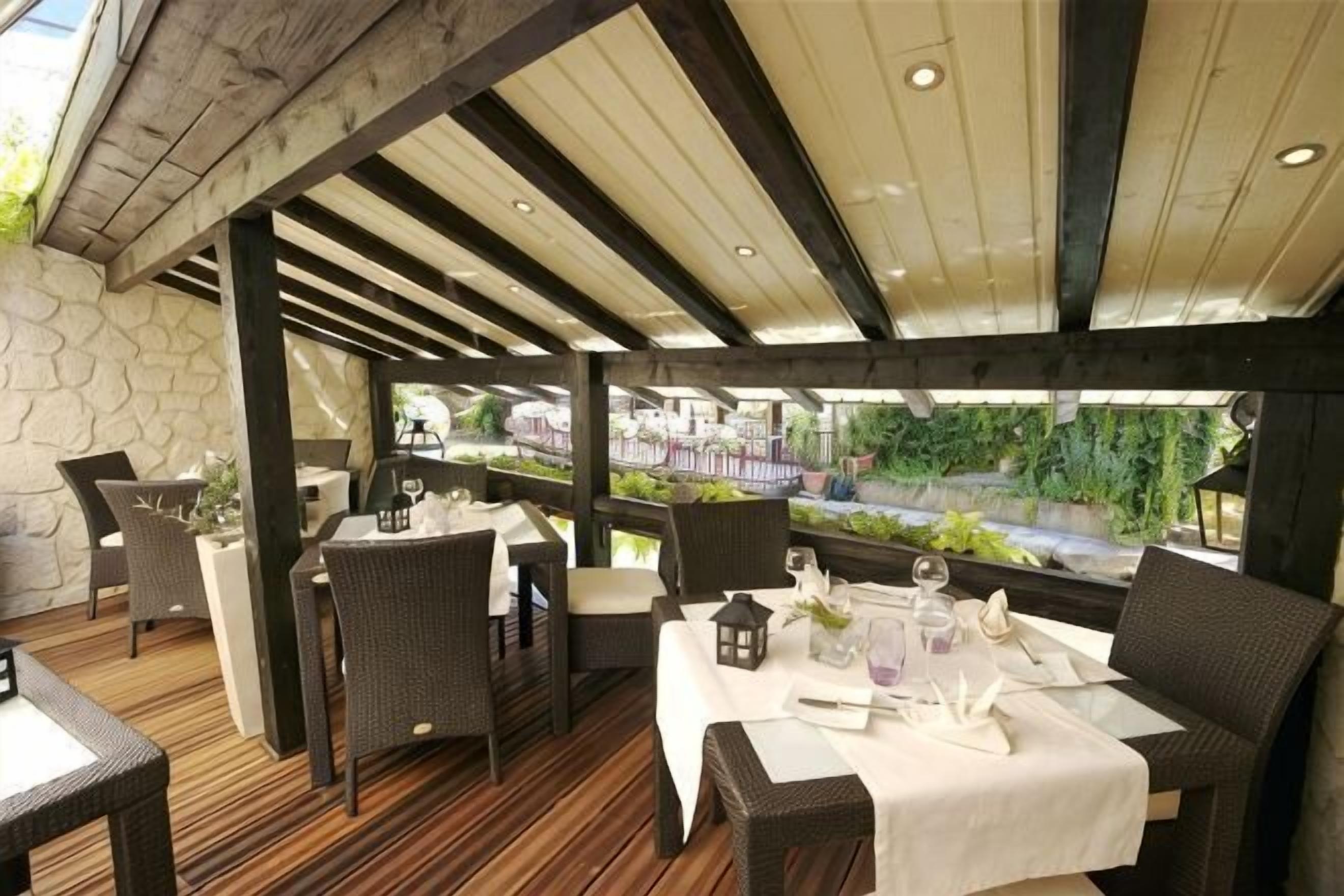 An outdoor dining area with tables set for two, covered by a wooden pergola, and a stone wall in the background.