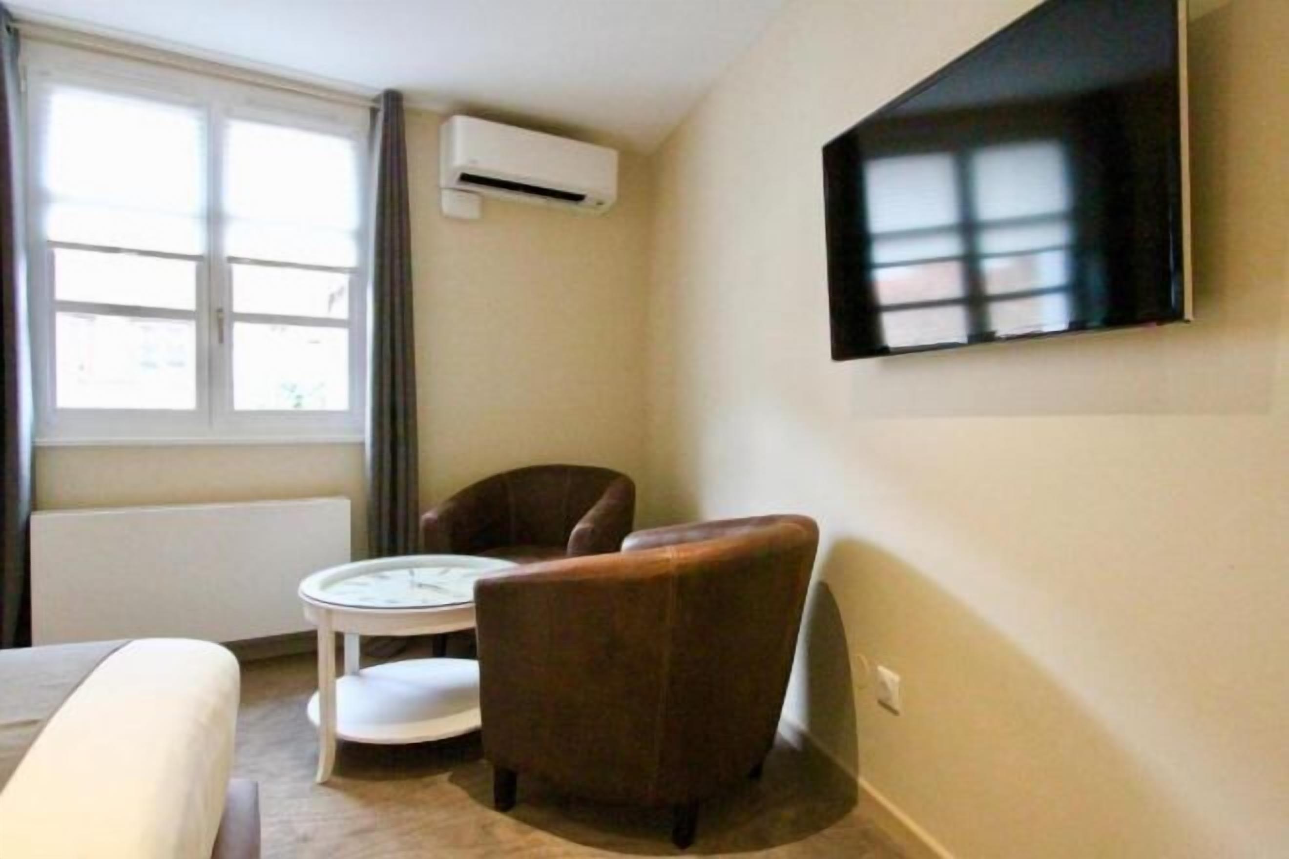 Una habitación con cama, una mesa redonda, un sillón marrón, un televisor de pantalla plana montado en la pared y una ventana con cortinas.