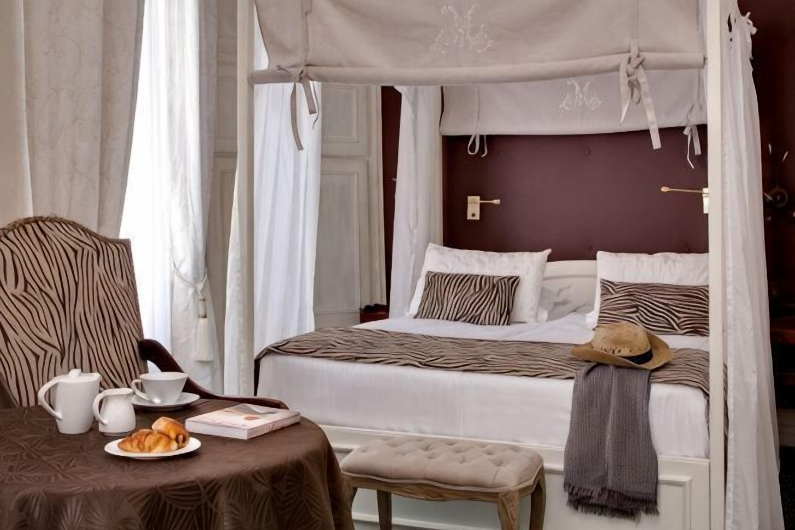 Une chambre à coucher avec un lit à baldaquin, une table de chevet sur laquelle se trouvent une tasse et une assiette, et un banc.