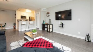 A modern living room with a flat-screen TV, a dining area with bar stools, a kitchen with stainless steel appliances, and a cozy seating area with a red and black checkered throw blanket.