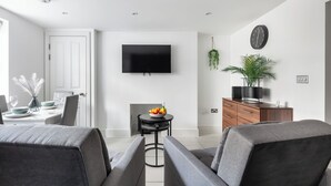 A modern living room with a flat-screen TV, grey sofas, a dining area with a table set, and a small side table with fruit.