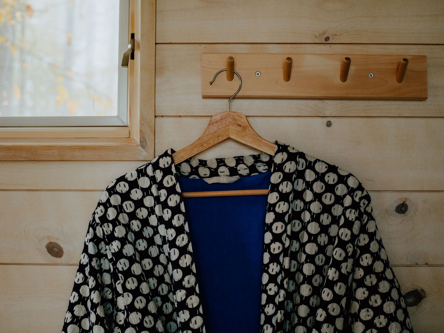 A wooden wall with a window and a coat rack with a blue shirt and a patterned jacket hanging on it.