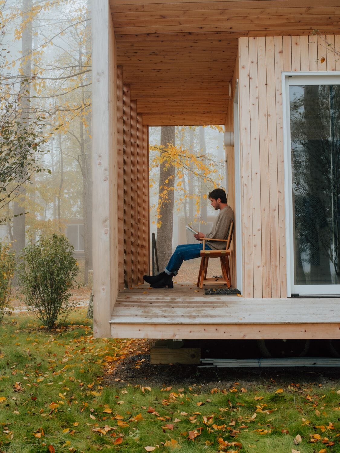 A person sitting on a wooden chair outside a wooden cabin with a glass door, surrounded by autumn leaves and trees.