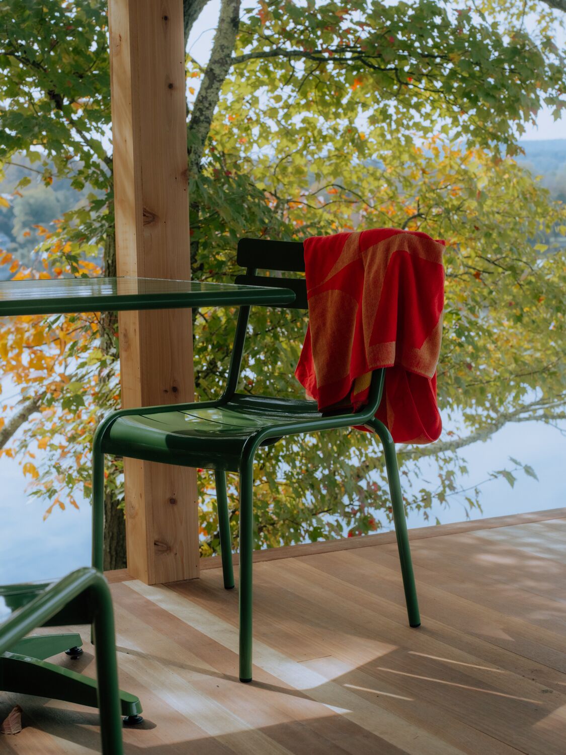 A green metal chair with a red towel draped over it, positioned on a wooden deck with a view of a lake and trees.