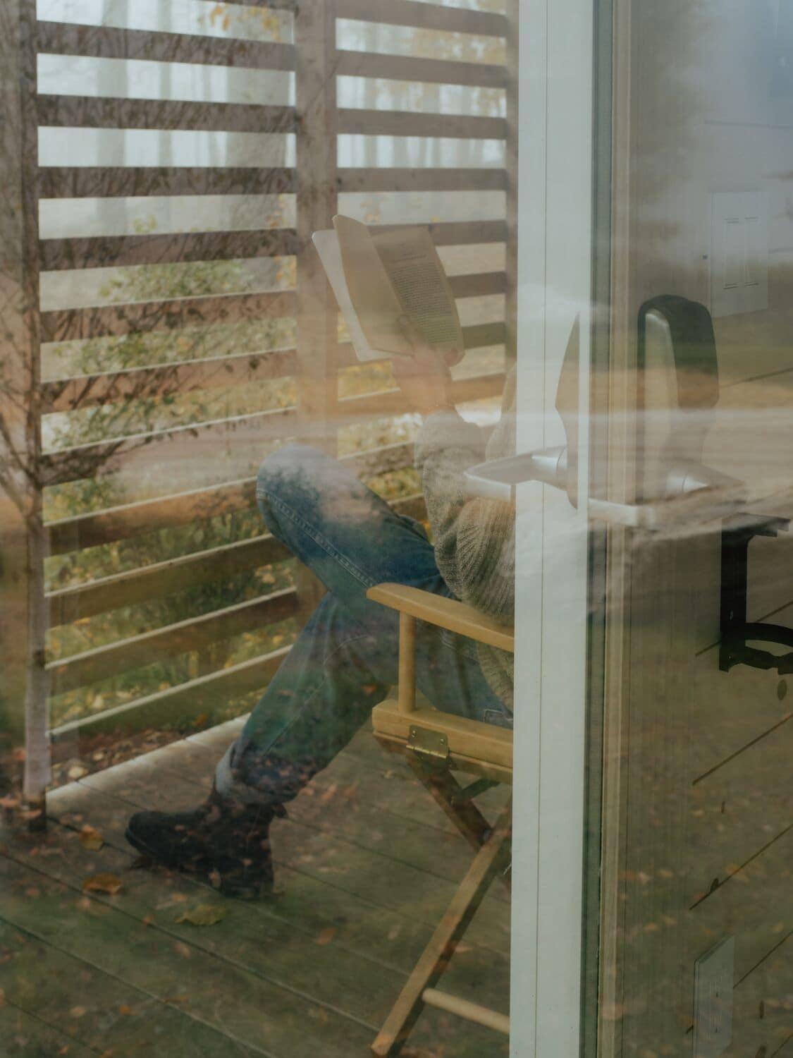 A wooden chair with a cushion, a wooden table, and a glass door reflecting an outdoor area with a wooden fence and greenery.