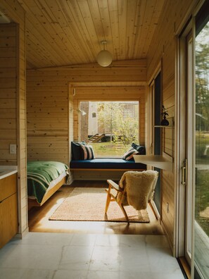 A cozy cabin room with a bed, a bench, a chair, and a large window offering a view of the outdoors. - Prospect Berkshires (Egremont)