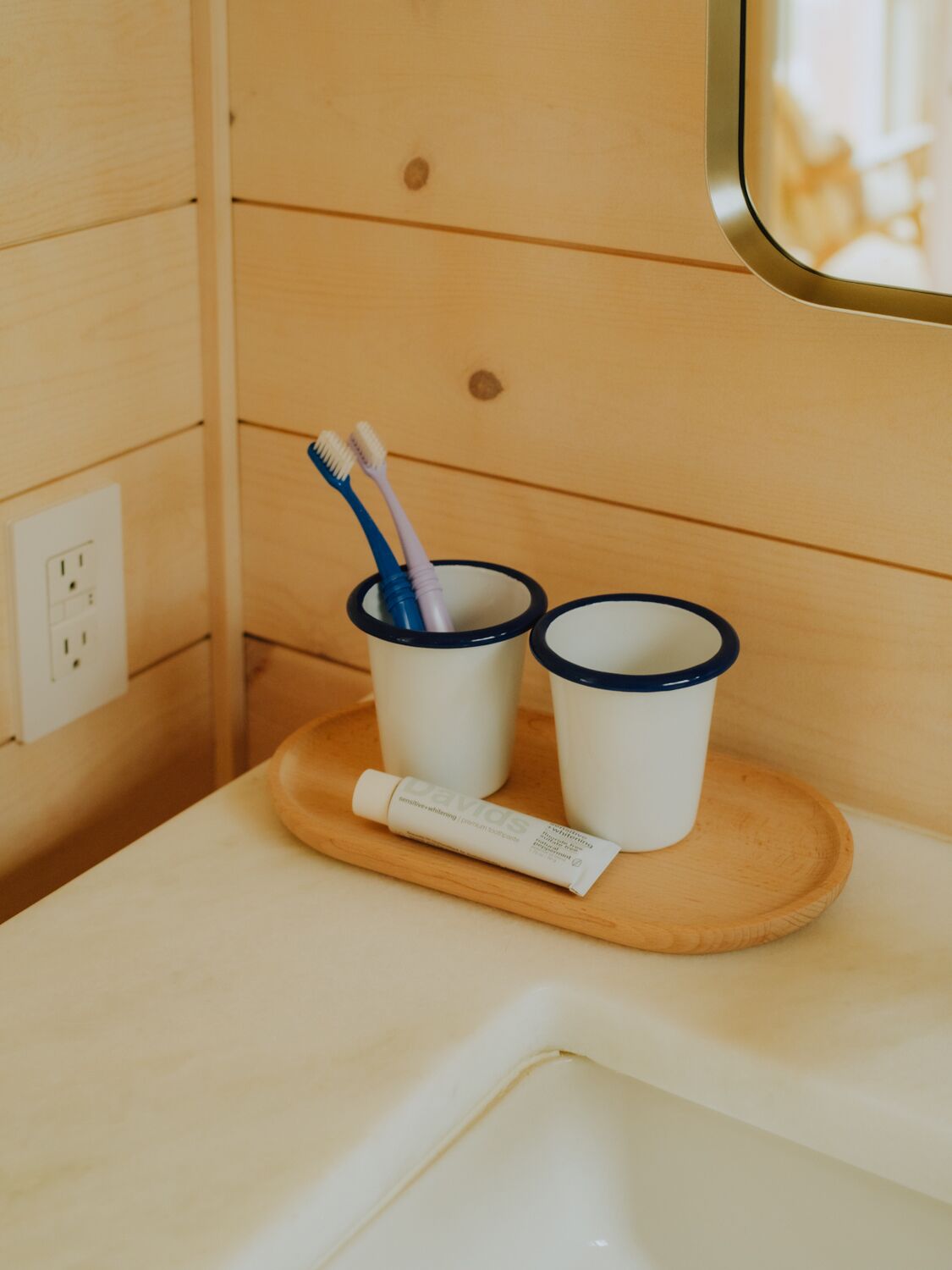 A wooden wall with a small window, a white sink, and two cups with toothbrushes.