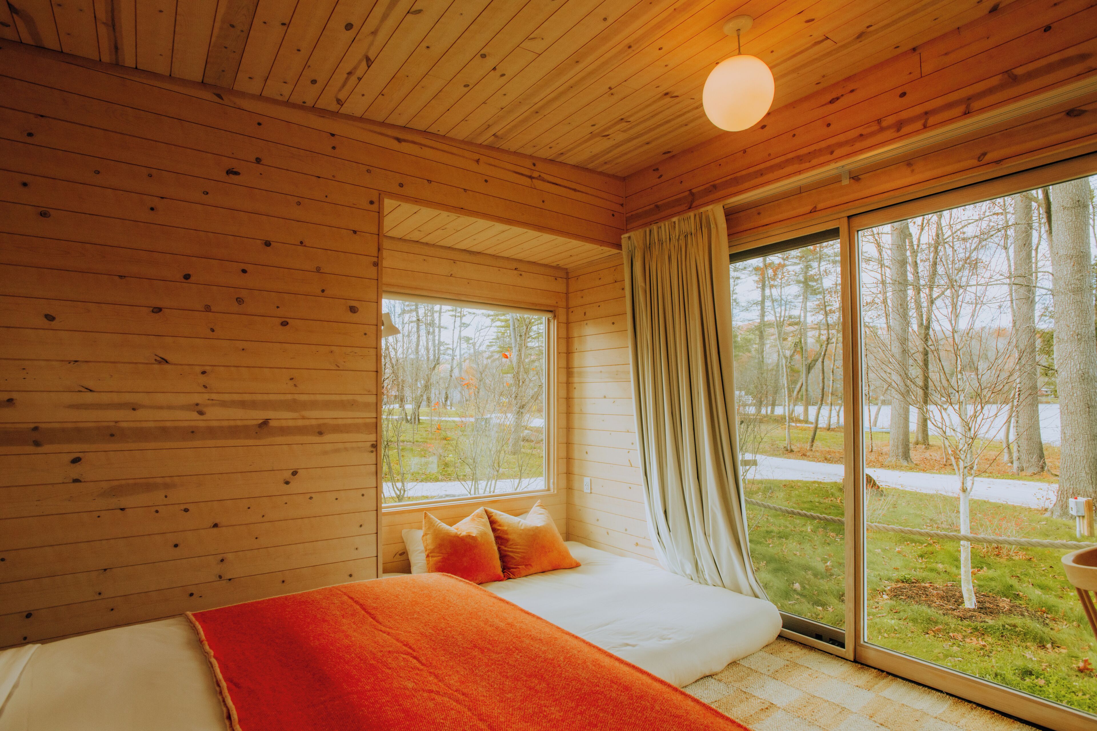A wooden cabin bedroom with a large window, a bed with orange bedding, and a view of trees and a path outside.
