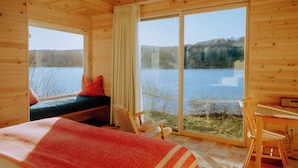 Una cabaña de madera con cama, una silla y una mesa junto a un ventanal grande con vista a un lago.