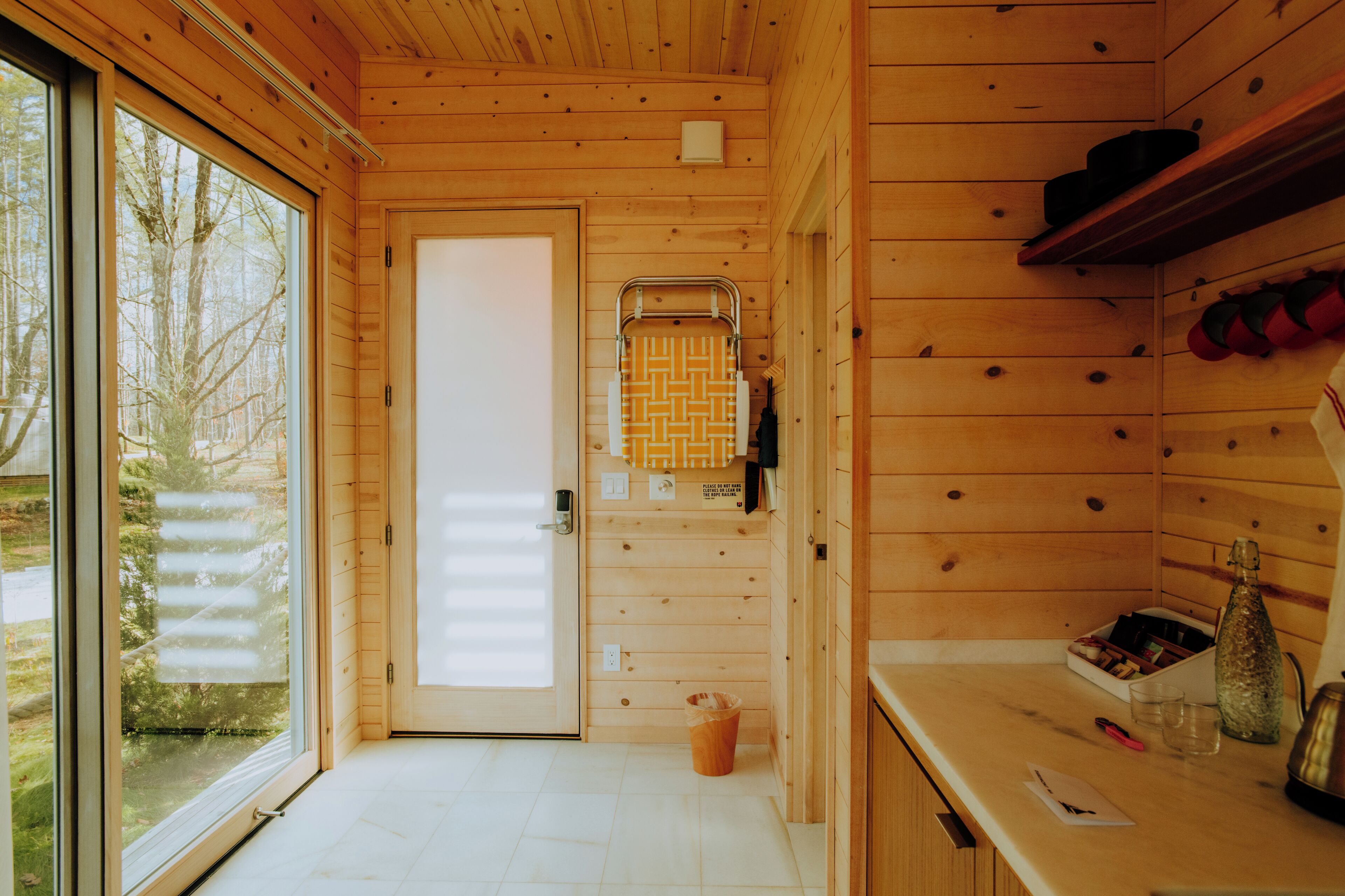 A wooden cabin interior with a large window, a door, a towel rack, and a counter with a bottle and glasses.