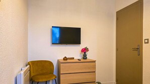 A room with a television mounted on the wall, a wooden dresser with drawers, a mustard-colored chair, and a door.