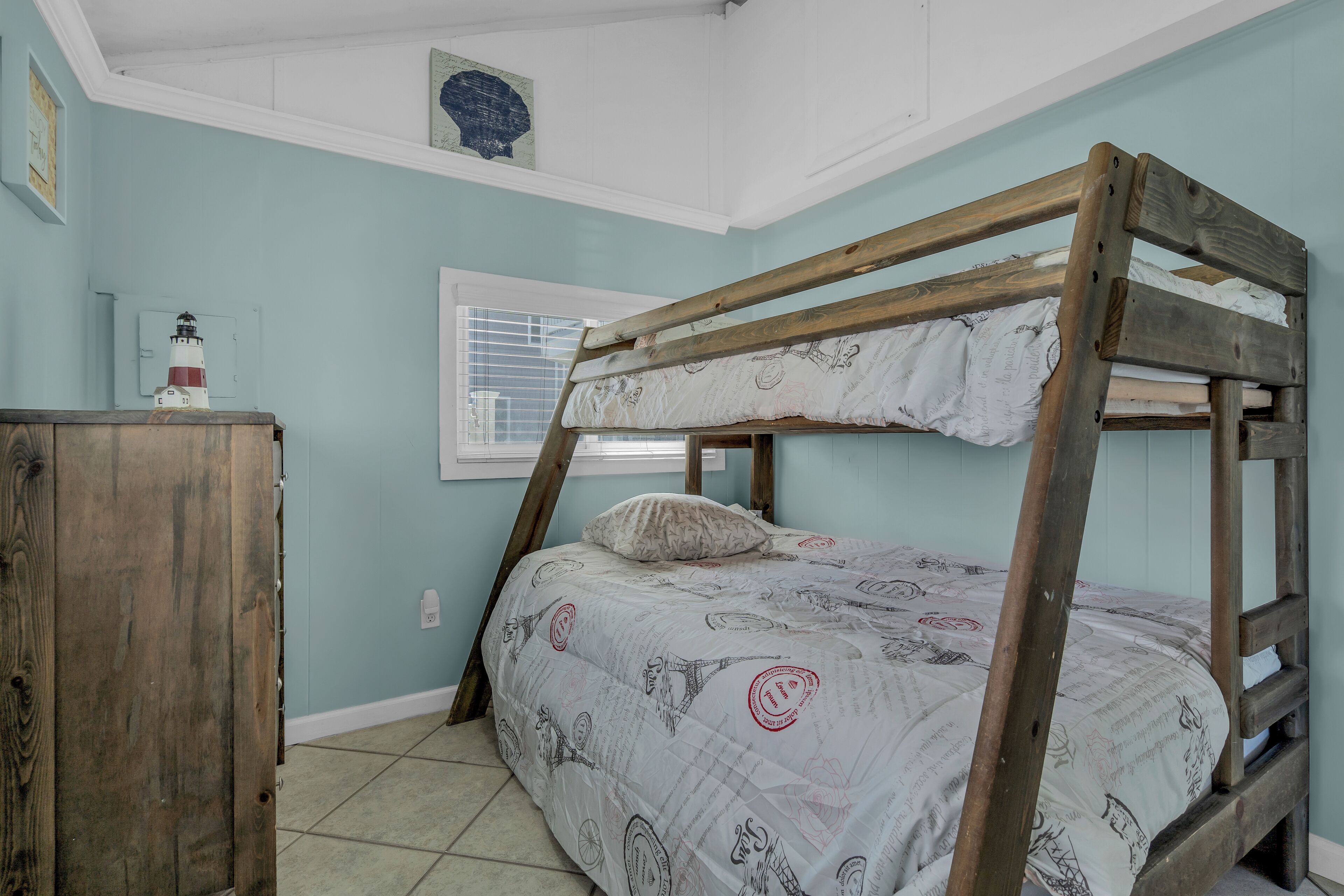 A bunk bed with a patterned bedspread, a wooden dresser, and a window with blinds.