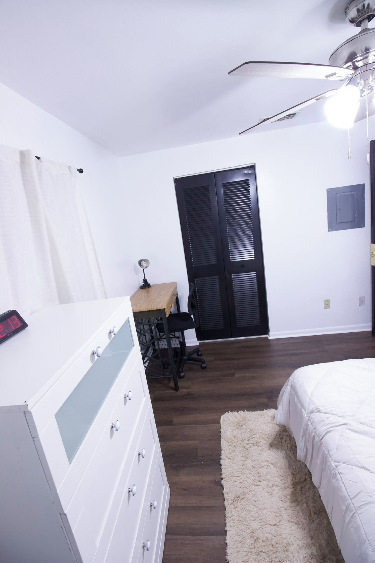 A bedroom with a bed, a dresser, a desk with a chair, a white curtain, a black door, and a ceiling fan.
