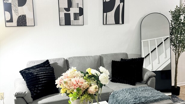 A living room with a grey sofa, a white coffee table, and a vase of flowers. There are abstract paintings on the wall and a mirror on the right. - Cozy 2-bedroom condo with WiFi, AC in quiet NW Gainesville, FL (Gainesville)