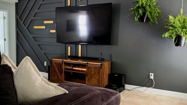 Una sala moderna con una pared oscura, un televisor de pantalla plana, un mueble de entretenimiento de madera, un sofá marrón y plantas colgantes.