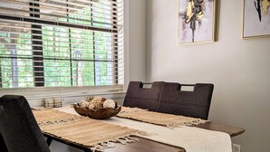 Un comedor con una mesa dispuesta para dos personas, una ventana con persianas y dos cuadros enmarcados en la pared.