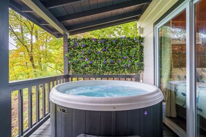 A hot tub on a wooden deck with a glass enclosure, surrounded by greenery and trees.