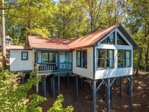 A two-story wooden house with a balcony, elevated on stilts, surrounded by trees.