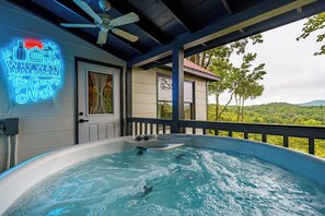 A hot tub with a neon sign, a ceiling fan, and a view of a house and trees. - The Crow’s Nest: Pet-friendly, Privacy, Mountain Views, Fire Pit & Deck! (Blue Ridge)