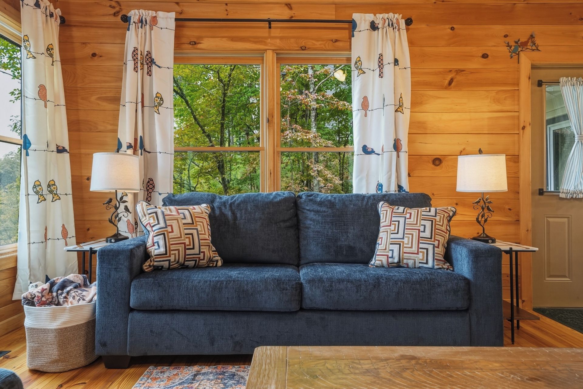 Une pièce chaleureuse avec un canapé bleu, des coussins à motifs, deux lampes et une fenêtre avec des rideaux.