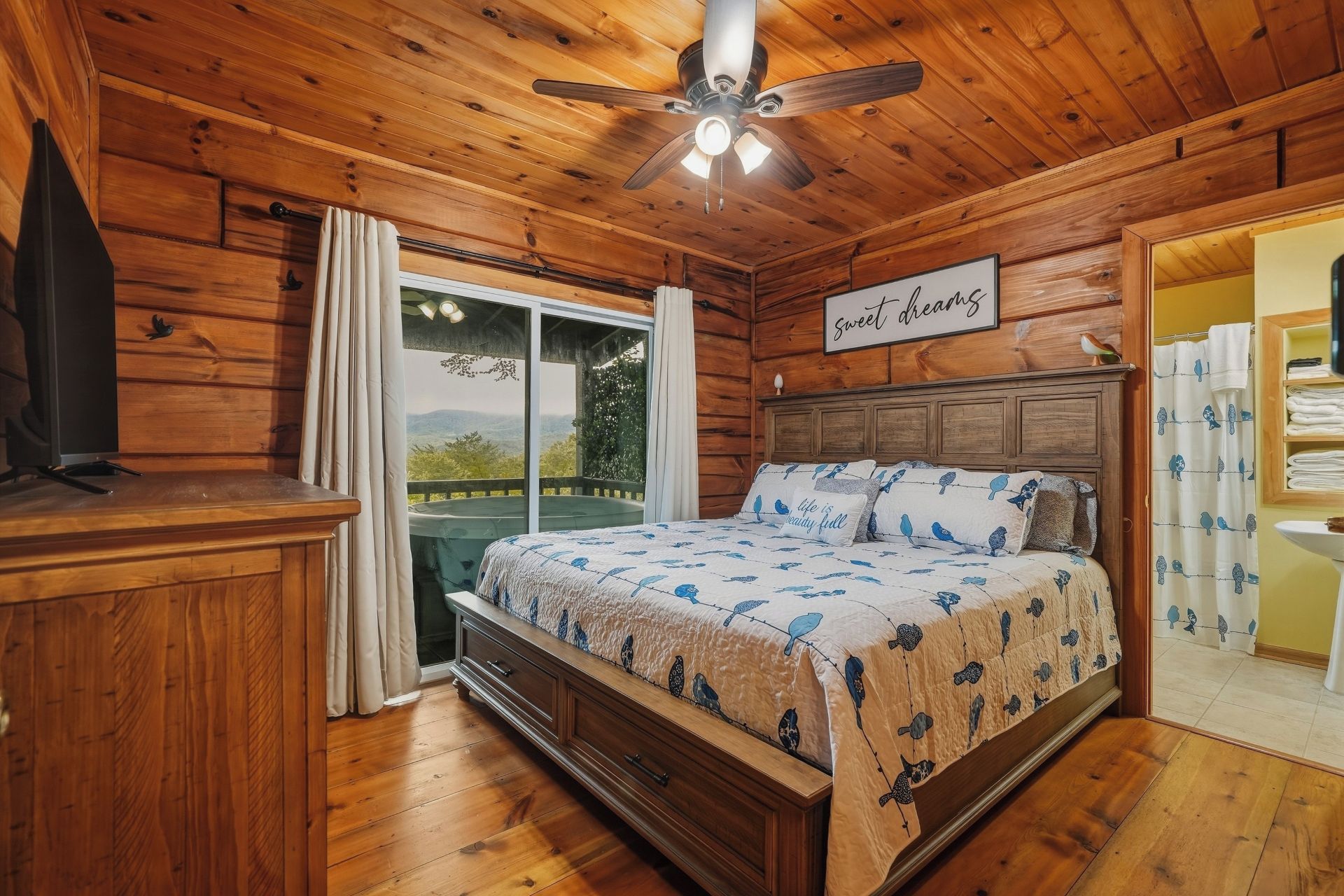 Une chambre avec un plafond en bois, un lit recouvert d’une couvre-lit à motifs d’oiseaux, un ventilateur de plafond et une vue sur l’extérieur grâce à une porte coulissante.