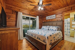 A bedroom with a wooden ceiling, a bed with a bird-patterned bedspread, a ceiling fan, and a view of the outdoors through a sliding door. - The Crow’s Nest: Pet-friendly, Privacy, Mountain Views, Fire Pit & Deck! (Blue Ridge)