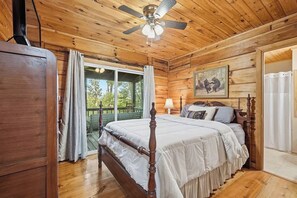 A wooden cabin bedroom with a bed, a ceiling fan, a window with curtains, and a painting on the wall.